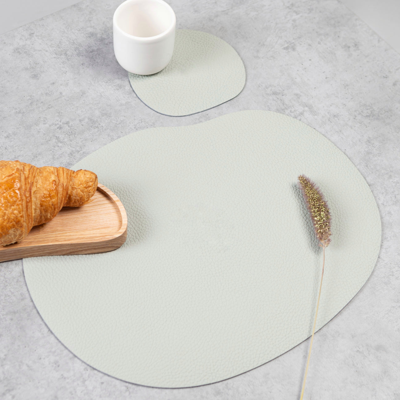 Light grey leather placemat and coaster with a croissant, smooth texture, and organic shape on a table.