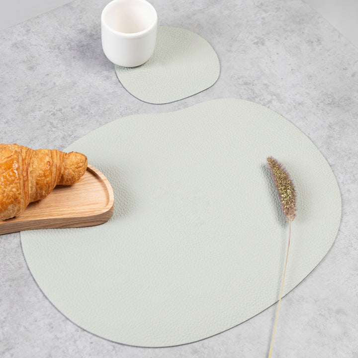Light grey leather placemat and coaster with a croissant, smooth texture, and organic shape on a table.