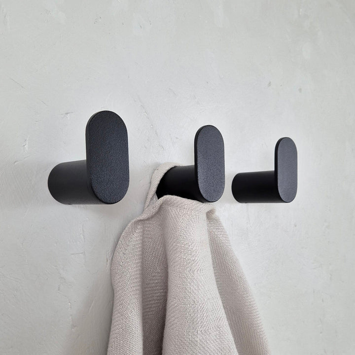 Minimalist wall hook in black, featuring clean lines and an oval shape, holding a towel against a textured wall.