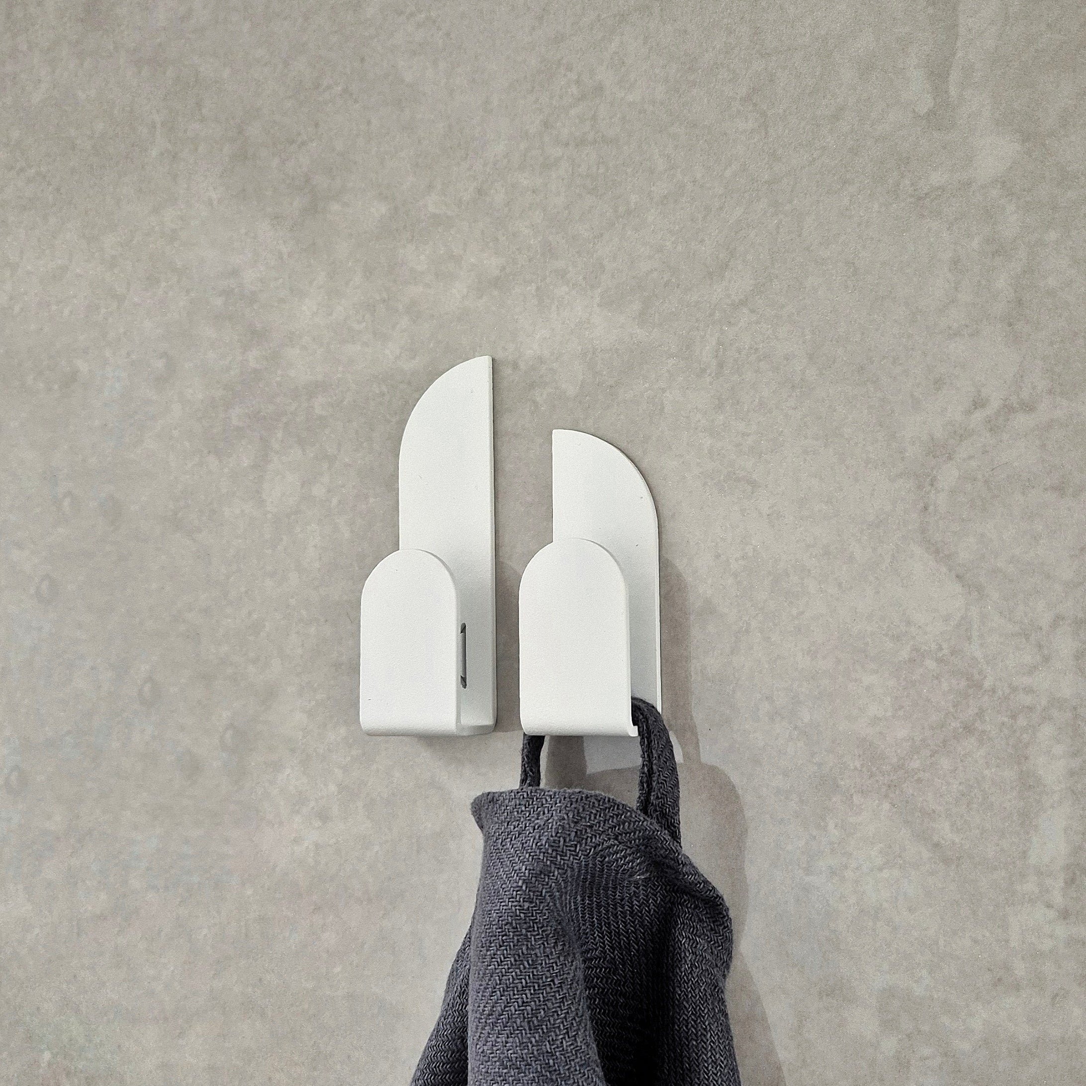 Adhesive wall hooks with a minimalist design holding a gray towel on a smooth wall.