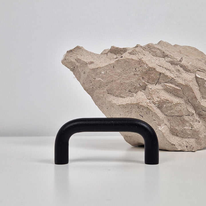 A matte black U-shaped cabinet pull handle displayed on a white surface in front of a textured beige stone prop against a light gray background.