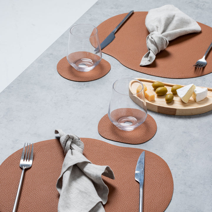 Terracotta leather placemat and coaster set on a dining table, featuring a minimalist design and wavy shapes.
