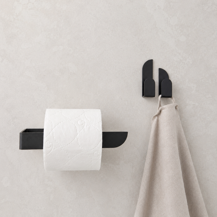 Adhesive black toilet paper holder mounted on a wall with a roll of toilet paper and a towel hanging beside it.