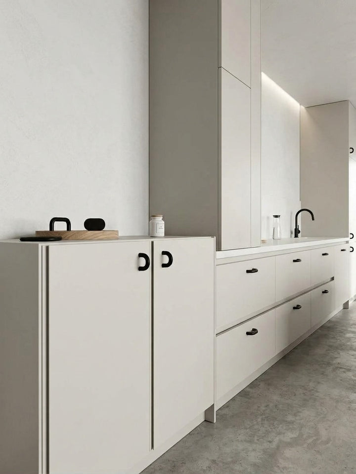 A modern minimalist kitchen cabinet set in warm off-white with contrasting black handles, set against a textured light gray wall and concrete-look floor.