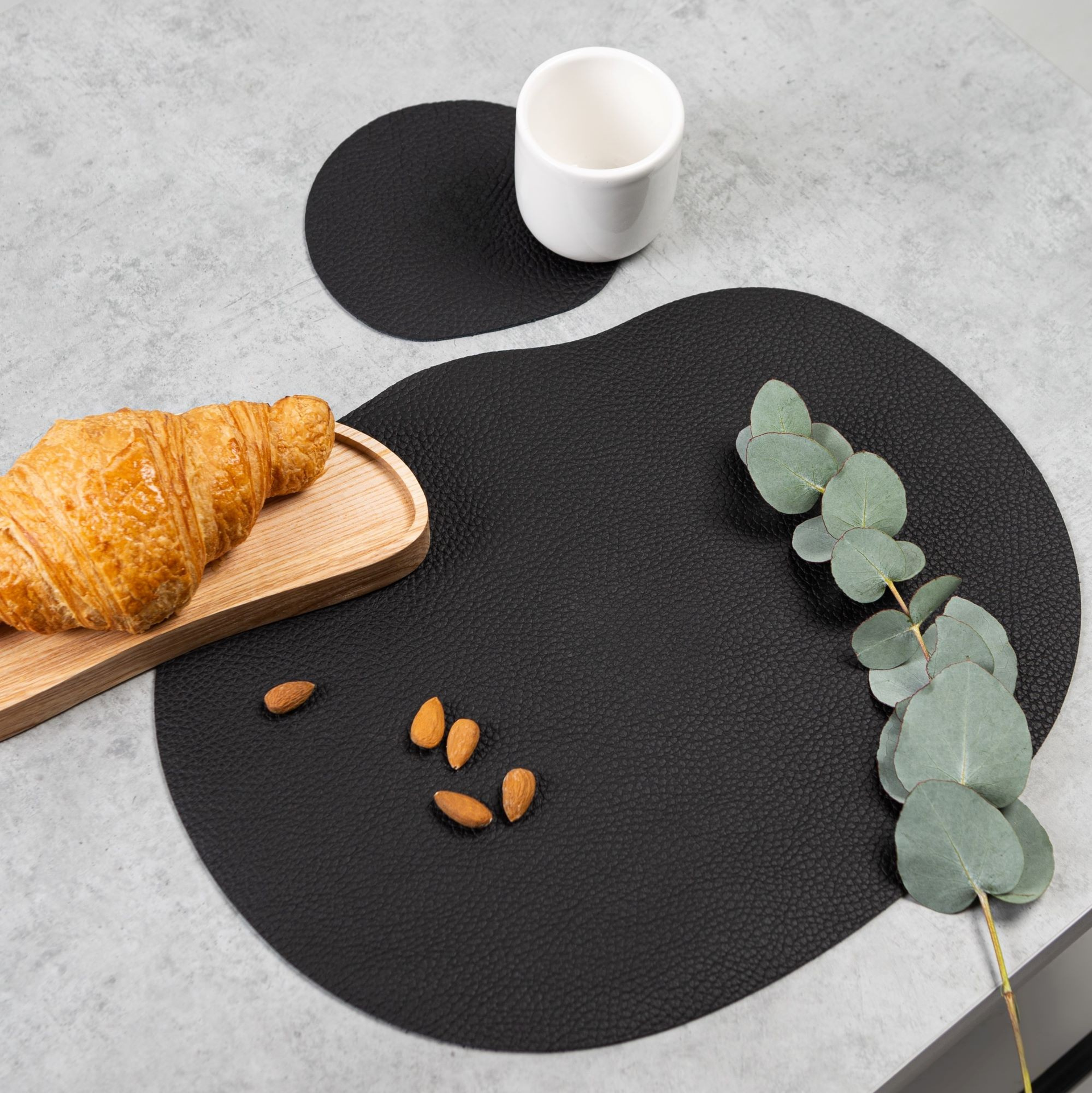 Black leather placemat and coaster MILLSTONES with croissant, almonds, and eucalyptus on table.